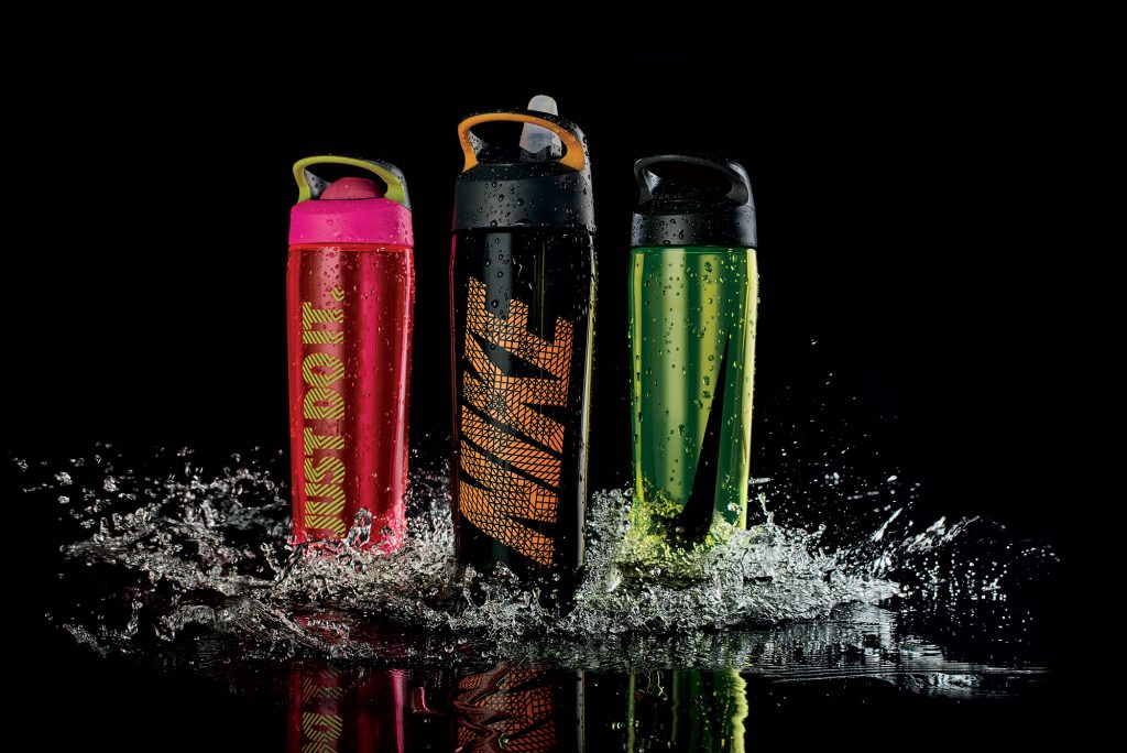 Three Nike water bottles in different colors surrounded by splashing water against a black background.