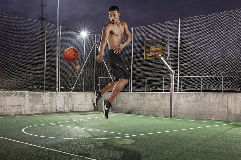 A shirtless man in shorts jumps while holding a basketball on an outdoor court at night.